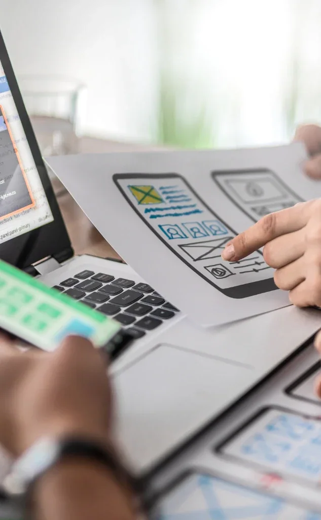 Fotografía de planificación de diseño web con un portátil, diseños de pantalla tipo wireframe y manos señalando un boceto de interfaz móvil.